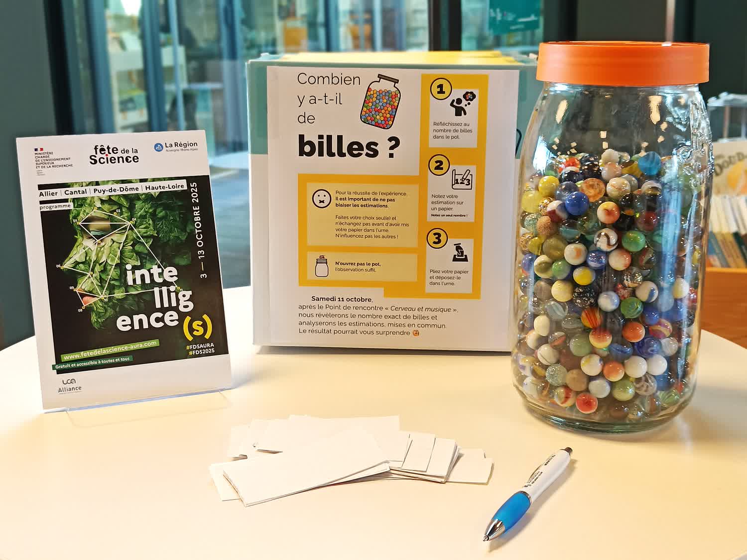 Photographie d'une table blanche, une boîte en carton avec un panneau expliquant la démarche, un grand pot en verre rempli à 80% de billes colorées, un flyer Fête de la science 2025, un tas de papiers découpés vierges avec un stylo.